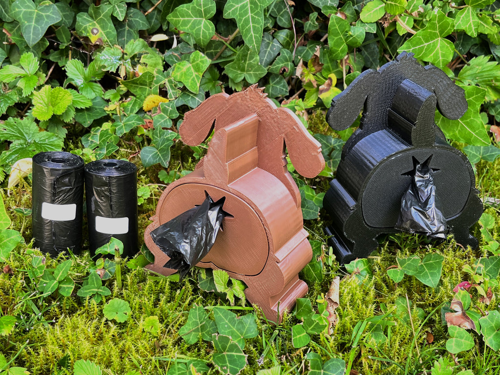 The Shitting Dog Kotbeutelspender in Braun und Schwarz nebeneinander im Garten, Kotbeutel kommt hinten heraus