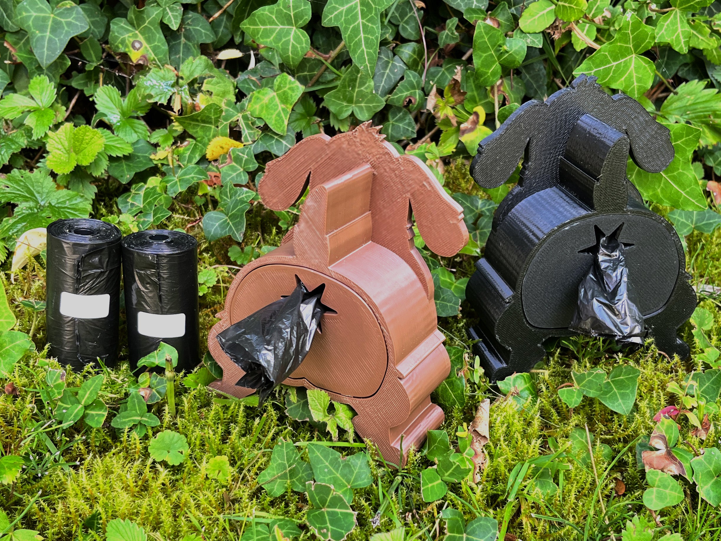 The Shitting Dog Kotbeutelspender in Braun und Schwarz nebeneinander im Garten, Kotbeutel kommt hinten heraus