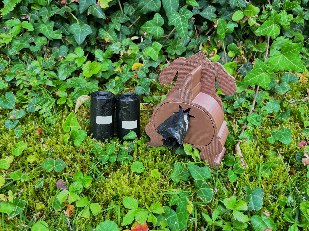 Brauner Shitting Dog Kotbeutelspender in Hundeform mit herausgezogenem Kotbeutel auf Moos