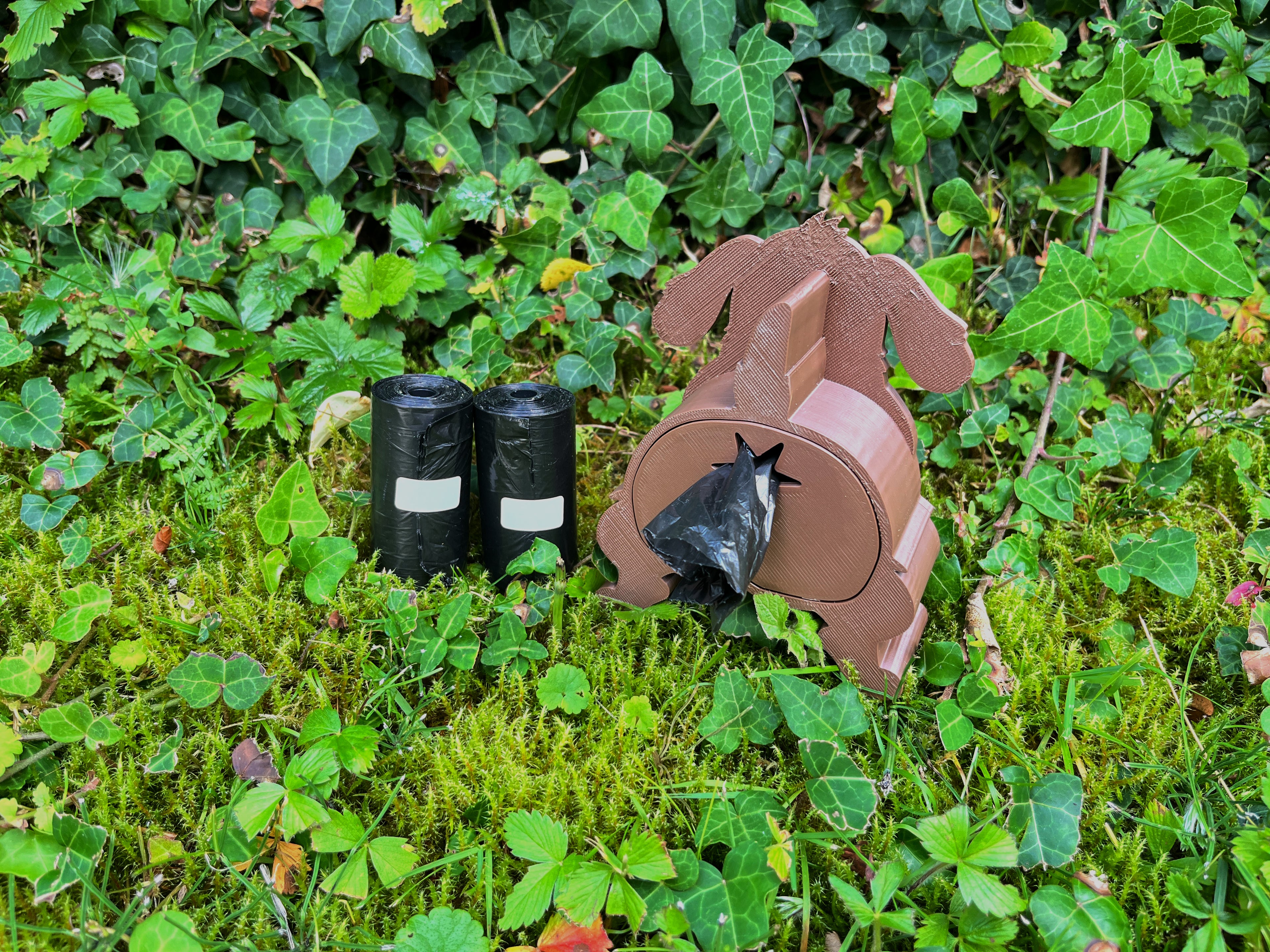 Brauner Shitting Dog Kotbeutelspender in Hundeform mit herausgezogenem Kotbeutel auf Moos